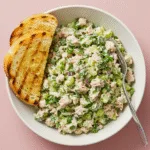 Close-up of a white bowl containing chunky tuna salad mixed with fresh herbs and diced vegetables, served with golden toasted bread slices and a fork on a pink surface