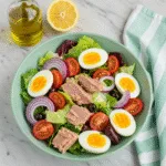 Colorful Niçoise salad in a mint green bowl containing mixed greens, chunks of tuna, halved hard-boiled eggs, cherry tomato wedges, and sliced red onion, with olive oil and lemon in the background Tuna in Water vs Oil